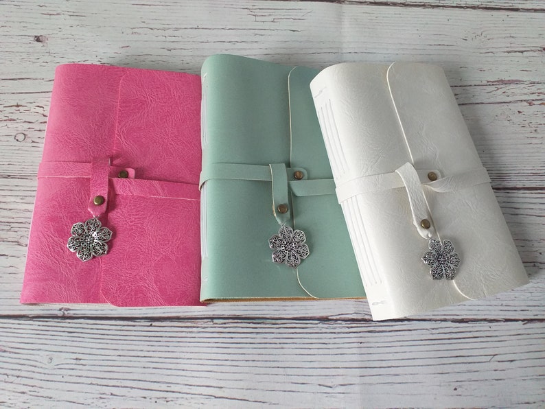 May include: Three leather journals in pink, mint green, and white. Each journal has a strap closure with a decorative silver flower charm. The journals are closed and appear to be new.