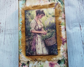 Puede incluir: Un diario de estilo vintage con un diseño floral y una cubierta de tela. La cubierta presenta una imagen de una mujer en un jardín recogiendo flores. El diario tiene un borde deshilachado y un borde de tela marrón alrededor de la imagen.