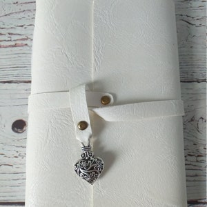 May include: A white journal with a textured cover and a strap closure. The strap is secured with a brass snap and features a decorative silver heart charm. The journal is closed and appears to be new.