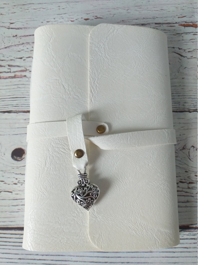 May include: A white journal with a textured cover and a strap closure. The strap is secured with a brass snap and features a decorative silver heart charm. The journal is closed and appears to be new.