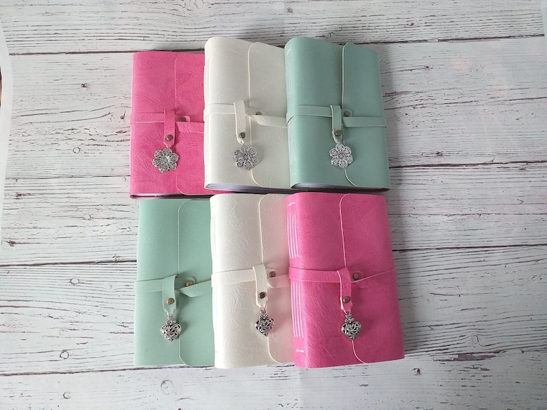 May include: A collection of six leather-bound journals in various colors: pink, white, and mint green. Each journal features a strap closure with a decorative silver charm. The journals are arranged on a light-colored wooden surface.