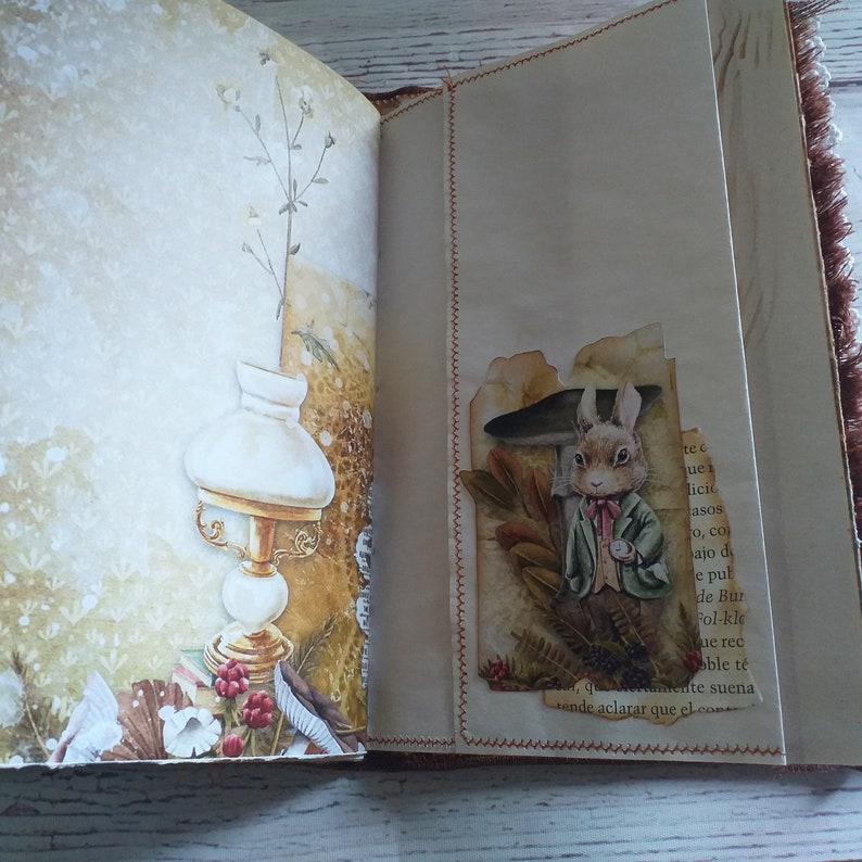 K&ouml;nnte beinhalten: Offenes Journal mit dekorativen Seiten. Eine Seite zeigt eine Vintage-Illustration einer Lampe und floralen Elementen. Die gegen&uuml;berliegende Seite hat eine Collage mit einem Kaninchen, das eine Jacke tr&auml;gt und eine Taschenuhr h&auml;lt. Das Journal hat einen braunen Einband und rote N&auml;hte.