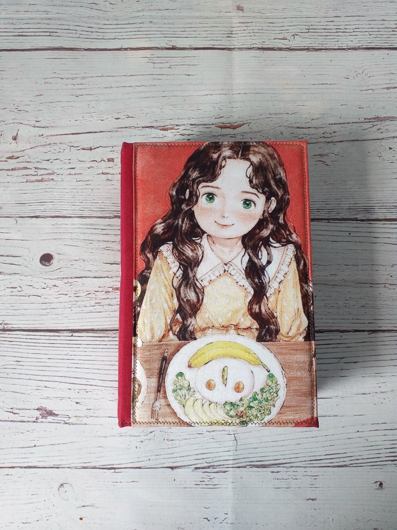 以下が含まれることがあります： 赤い背表紙と前面にカラフルなイラストが描かれた布張りのジャーナル。イラストは、長い茶色の髪と黄色のドレスを着た若い女の子が、食べ物のプレートのあるテーブルに座っている様子を描いています。