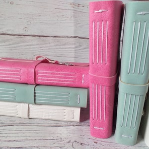 May include: A stack of handmade leather journals in various colors: pink, seafoam green, and white. Each journal features a stitched spine and a leather strap closure. The journals are displayed against a light wood background.