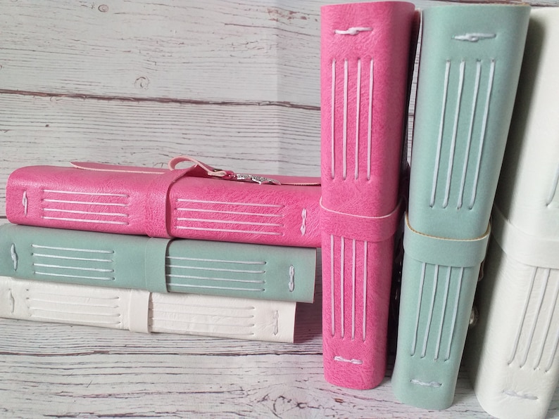 May include: A stack of handmade leather journals in various colors: pink, seafoam green, and white. Each journal features a stitched spine and a leather strap closure. The journals are displayed against a light wood background.