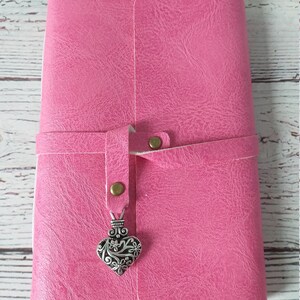 May include: A pink journal with a textured cover and a strap closure. The strap is secured with a brass-colored snap. A decorative silver heart charm hangs from the strap, adding a touch of elegance.