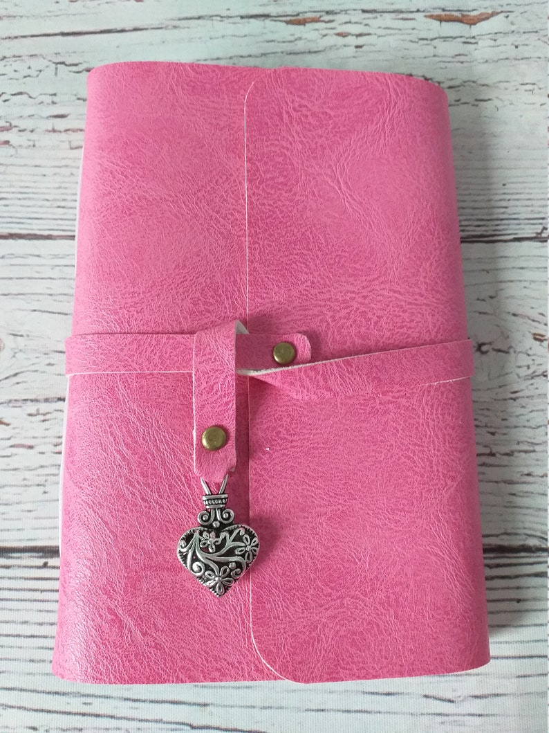 May include: A pink journal with a textured cover and a strap closure. The strap is secured with a brass-colored snap. A decorative silver heart charm hangs from the strap, adding a touch of elegance.
