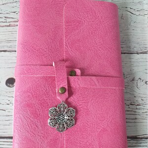 May include: A pink journal with a textured cover and a strap closure. The strap is secured with a bronze-colored button. A decorative silver-colored flower charm hangs from the strap. The journal is closed.