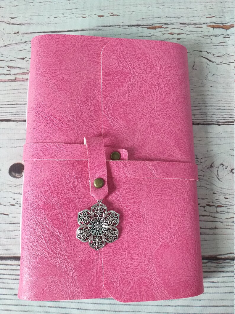 May include: A pink journal with a textured cover and a strap closure. The strap is secured with a bronze-colored button. A decorative silver-colored flower charm hangs from the strap. The journal is closed.