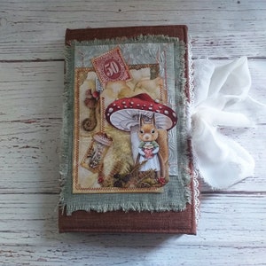 K&ouml;nnte beinhalten: Ein braunes Notizbuch mit einem skurrilen Design, das ein Eichh&ouml;rnchen unter einem roten Pilz zeigt. Das Cover hat eine Collage von Illustrationen im Vintage-Stil, eine Briefmarke und ein wei&szlig;es Band. Die Kanten sind mit ausgefranstem gr&uuml;nem Stoff beschnitten.
