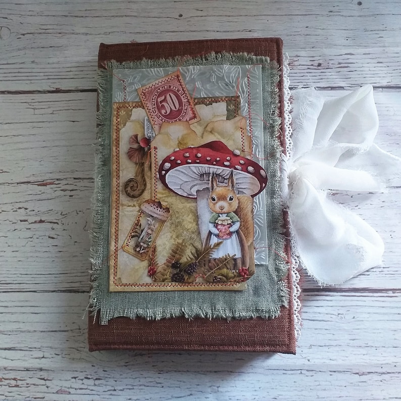 K&ouml;nnte beinhalten: Ein braunes Notizbuch mit einem skurrilen Design, das ein Eichh&ouml;rnchen unter einem roten Pilz zeigt. Das Cover hat eine Collage von Illustrationen im Vintage-Stil, eine Briefmarke und ein wei&szlig;es Band. Die Kanten sind mit ausgefranstem gr&uuml;nem Stoff beschnitten.