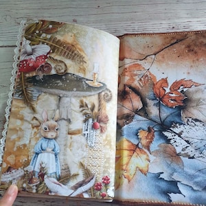 K&ouml;nnte beinhalten: Aufgeschlagenes Journal mit einer skurrilen Illustration mit einem Kaninchen in einem blauen Kleid, das eine Teetasse, Pilze und Herbstbl&auml;tter h&auml;lt. Die Seiten haben ein Vintage-Aussehen mit Spitzenbesatz.