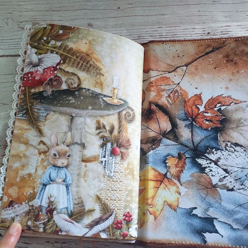 K&ouml;nnte beinhalten: Aufgeschlagenes Journal mit einer skurrilen Illustration mit einem Kaninchen in einem blauen Kleid, das eine Teetasse, Pilze und Herbstbl&auml;tter h&auml;lt. Die Seiten haben ein Vintage-Aussehen mit Spitzenbesatz.