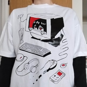 May include: White t-shirt with a black and white illustration of a person looking out of a computer monitor. The illustration also includes a keyboard, headphones, a mouse, and a handheld video game console.
