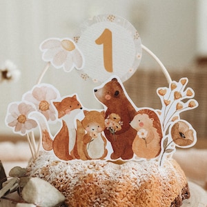 May include: A woodland-themed cake topper with a brown bear, fox, hedgehog, and a number one. The topper is on a cake with white powdered sugar. The cake is on a wooden cake stand.
