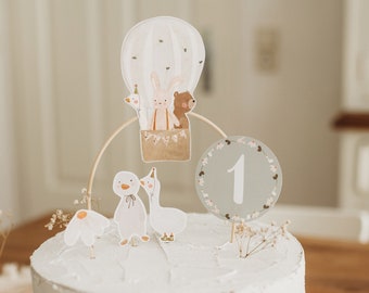 May include: A white cake with a white frosting and a hot air balloon topper with animals. The balloon is decorated with brown, white, and beige. The cake is decorated with a white number one on a gray circle with green leaves around it. The cake is on a wooden table with a brown and white blanket.