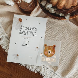 Peut inclure: Une feuille d'instructions blanche avec le texte "B&uuml;gelbild ANLEITUNG" et un symbole de fer &agrave; repasser, ainsi qu'un petit carr&eacute; avec un dessin d'ours et le texte "ICH BIN EINS". Les articles sont sur un tissu beige avec une bordure &agrave; franges.