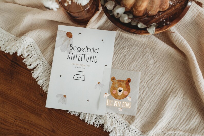 Peut inclure: Une feuille d'instructions blanche avec le texte "B&uuml;gelbild ANLEITUNG" et un symbole de fer &agrave; repasser, ainsi qu'un petit carr&eacute; avec un dessin d'ours et le texte "ICH BIN EINS". Les articles sont sur un tissu beige avec une bordure &agrave; franges.