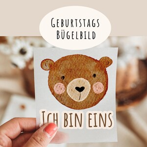 Könnte beinhalten: Ein braunes Bügelbild mit einem Comic-Bärenkopf und dem Text "Ich bin eins". Das Bügelbild befindet sich auf einer durchsichtigen Plastikfolie.