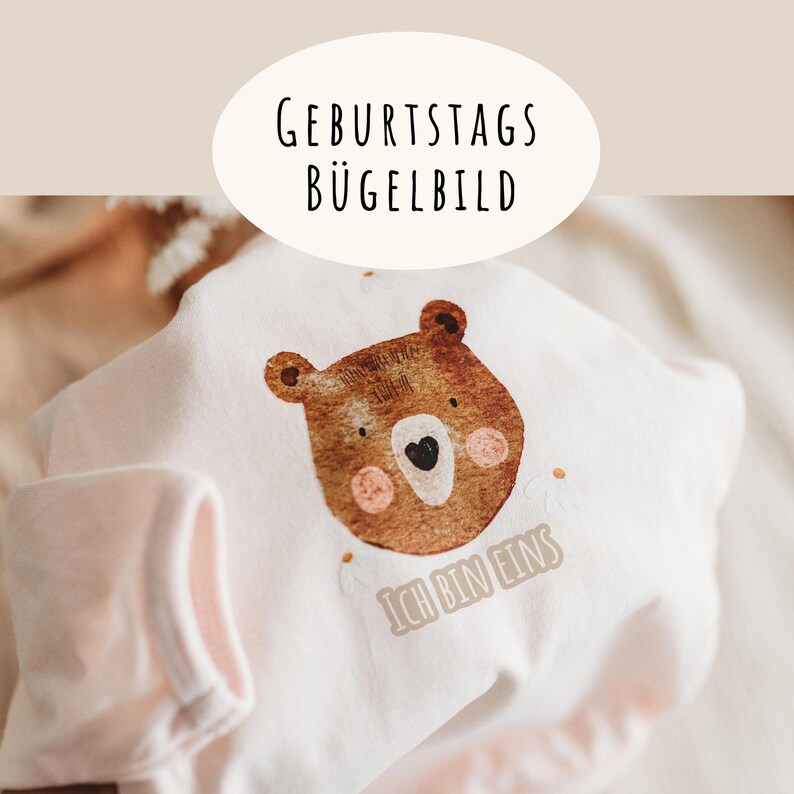 Peut inclure: Un body de b&eacute;b&eacute; rose clair avec un visage d'ours brun, des accents roses et le texte "ICH BIN EINS". Le texte "GEBURTSTAGS B&Uuml;GELBILD" est au-dessus de l'ours en noir. Le body est pli&eacute;, montrant une partie de la manche.