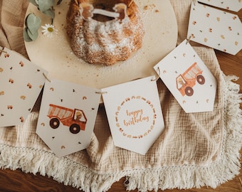 May include: A brown and white birthday banner with a truck design and the words "happy birthday" in a circle. The banner is hanging on a beige fabric with a fringe trim.
