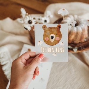 Peut inclure: Une carte avec une illustration d'ours brun et le texte "Ich bin eins". La carte est tenue devant un fond flou avec un g&acirc;teau et des fleurs, avec une table en bois en arri&egrave;re-plan.