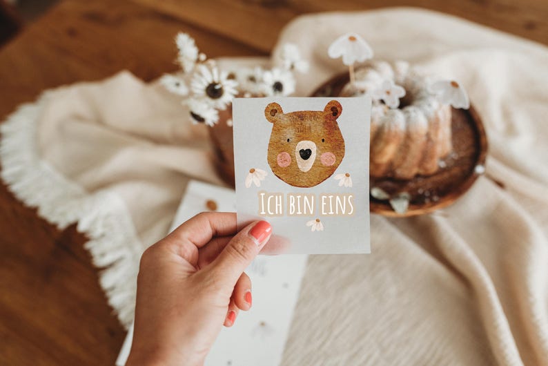 Peut inclure: Une carte avec une illustration d'ours brun et le texte "Ich bin eins". La carte est tenue devant un fond flou avec un g&acirc;teau et des fleurs, avec une table en bois en arri&egrave;re-plan.
