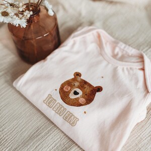 Peut inclure: Un body de b&eacute;b&eacute; rose clair avec un dessin de t&ecirc;te d'ours brun et le texte "Ich bin eins". Le body est pli&eacute; sur un tissu beige. Un vase marron avec des fleurs s&eacute;ch&eacute;es est en arri&egrave;re-plan.