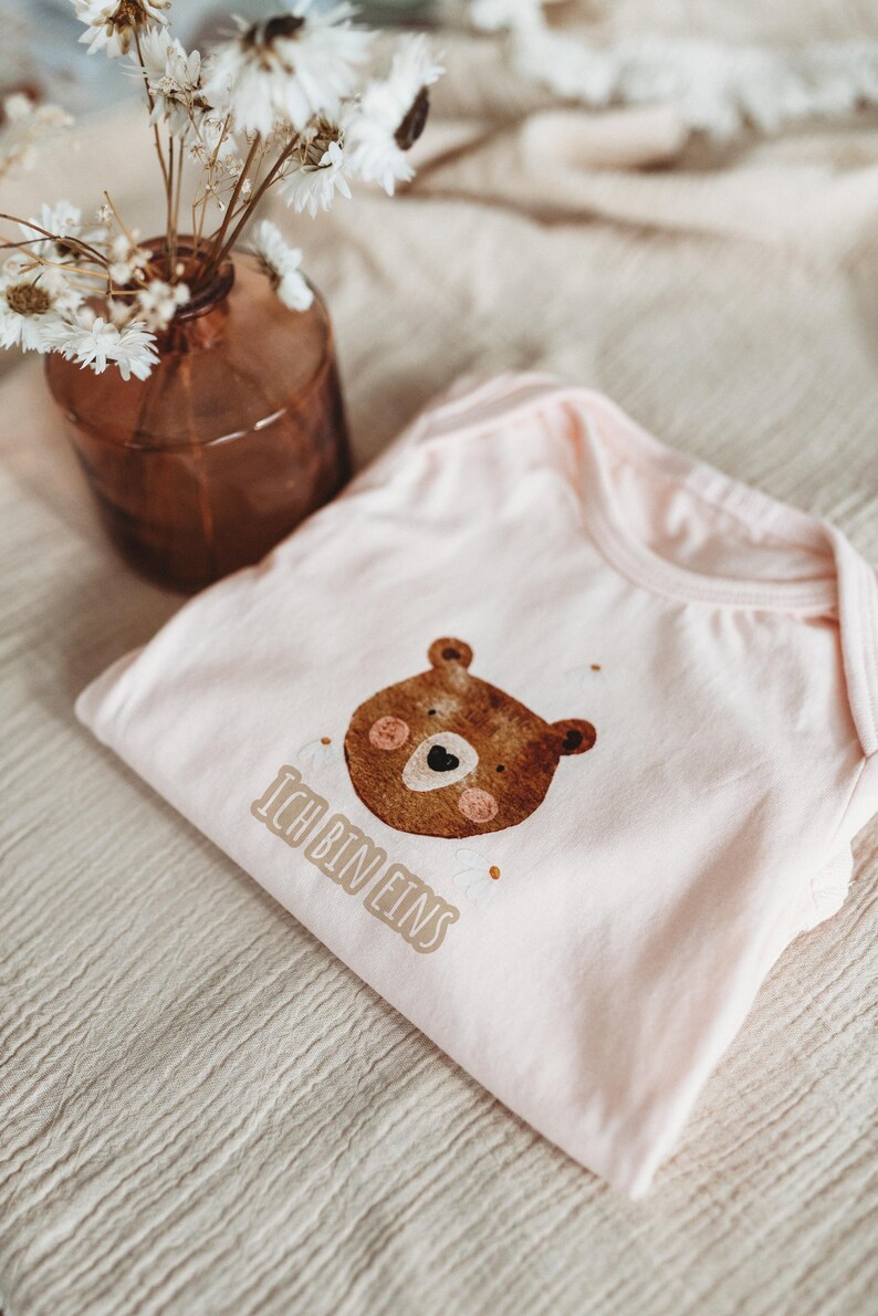 Peut inclure: Un body de b&eacute;b&eacute; rose clair avec un dessin de t&ecirc;te d'ours brun et le texte "Ich bin eins". Le body est pli&eacute; sur un tissu beige. Un vase marron avec des fleurs s&eacute;ch&eacute;es est en arri&egrave;re-plan.