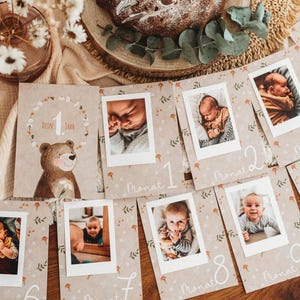 Pu&ograve; includere: Un set di biglietti per tappe fondamentali del bambino con foto, con un'illustrazione di orso e il testo "Dein 1 Jahr". Le carte sono disposte su una superficie di legno, con una torta e fiori sullo sfondo. Ogni carta ha una foto del bambino.