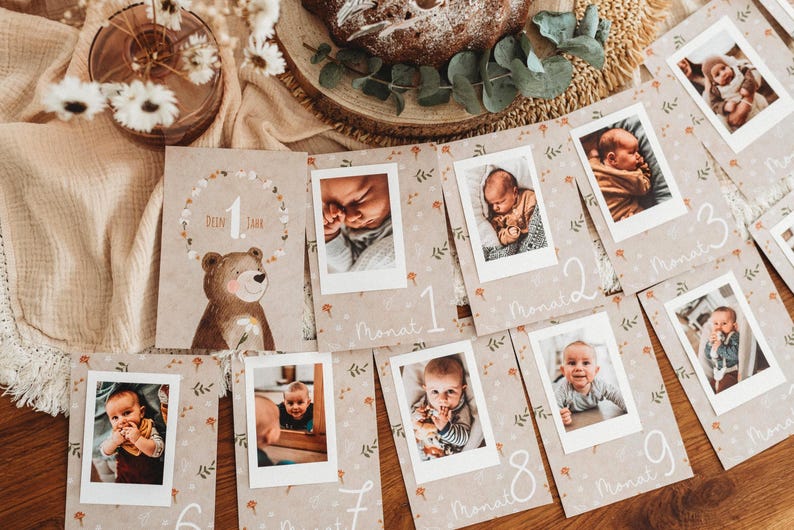 Pu&ograve; includere: Un set di biglietti per tappe fondamentali del bambino con foto, con un'illustrazione di orso e il testo "Dein 1 Jahr". Le carte sono disposte su una superficie di legno, con una torta e fiori sullo sfondo. Ogni carta ha una foto del bambino.