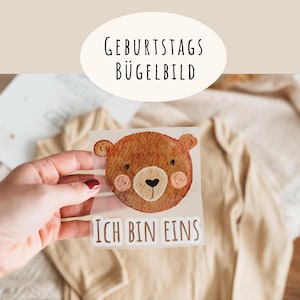 May include: A brown bear iron-on transfer with the text "Ich bin eins" and "Geburtstags Bügelbild". The bear has pink cheeks and a black heart-shaped nose. The transfer is held over a beige onesie.