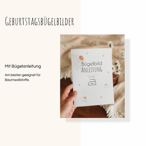 Peut inclure: Un livret blanc intitul&eacute; "Geburtstagsb&uuml;gelbilder" avec les mots "Mit B&uuml;gelanleitung" et "Am besten geeignet f&uuml;r Baumwollstoffe." Le livret pr&eacute;sente un motif de marguerite et un symbole de fer &agrave; repasser. Une main tient le livret.