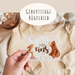 Könnte beinhalten: Beiger Baby-Body mit einem Bügelbild mit einem braunen Bären und einem Hasen, mit dem Text "Ich bin eins". Das Bild enthält auch den Text "Geburtstags Bügelbild" in einem weißen Oval.