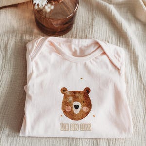 Peut inclure: Un body de b&eacute;b&eacute; rose clair avec un dessin de visage d'ours brun. L'ours a un nez en forme de c&oelig;ur et des joues roses. Le texte "ICH BIN EINS" est imprim&eacute; en dessous. Le body est pli&eacute; sur un tissu beige.