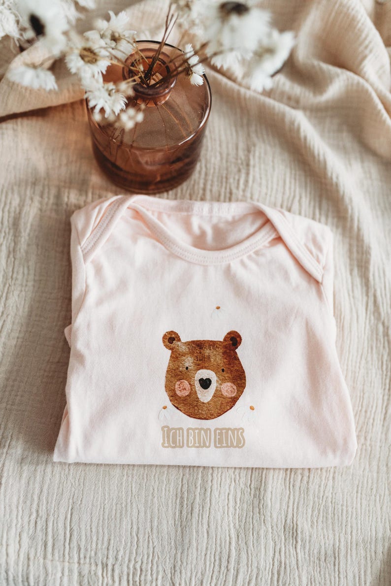 Peut inclure: Un body de b&eacute;b&eacute; rose clair avec un dessin de visage d'ours brun. L'ours a un nez en forme de c&oelig;ur et des joues roses. Le texte "ICH BIN EINS" est imprim&eacute; en dessous. Le body est pli&eacute; sur un tissu beige.