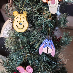 May include: Four hanging ornaments shaped like cartoon characters: a yellow bear, an orange and black striped tiger, a blue and pink donkey, and a pink piglet. Each ornament features a gold string loop for hanging on a green artificial tree branch.