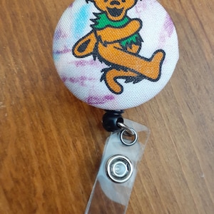 Puede incluir: Un portatarjetas redondo con un diseño de oso bailarín en naranja y negro, con un cuello verde, sobre un fondo teñido. El portatarjetas tiene una correa de plástico transparente y un cable retráctil negro.