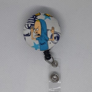 May include: A white, blue, and yellow button with a cartoon character wearing a blue hoodie and holding up his hands. The button is attached to a clear plastic retractable badge holder with a black spring.
