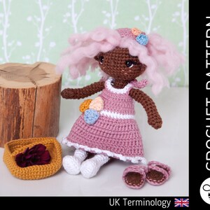 Könnte beinhalten: Häkelanleitung für eine Puppe mit rosa Haaren, einem rosa Kleid und weißen Schuhen. Die Puppe sitzt auf einem Baumstumpf mit einem kleinen Korb daneben. Die Anleitung ist in britischer Terminologie geschrieben.