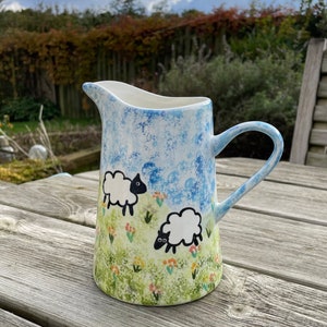 May include: A hand-painted ceramic jug with a sheep motif. The jug has a blue and white sky design at the top, with a green field and two sheep. The handle and rim are light blue. The jug is on a wooden table.