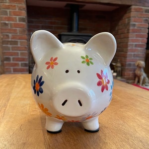May include: A white ceramic piggy bank decorated with colourful flowers in orange, blue, and pink. The piggy bank has a classic pig shape with four black feet and a slot on top for inserting coins. A charming decorative item.