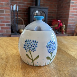 May include: A white ceramic sugar bowl with a light blue lid and a small handle. The bowl is decorated with blue floral designs and green grass accents. The bowl is approximately 13 cm tall.