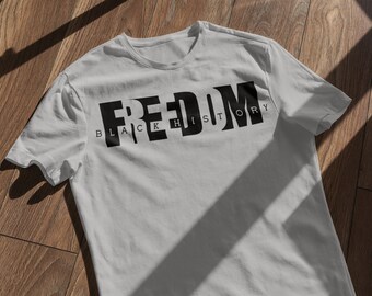 May include: A light gray t-shirt with the word "FREEDOM" in large, bold, black letters. Below the word "FREEDOM" is the text "BLACK HISTORY" in smaller, black letters. The shirt is made of a soft material.