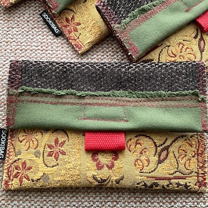 May include: Close-up of several fabric pouches with the text "SOOKIBAG" visible on the side. The pouches feature a variety of textures and colours, including olive green, gold floral, and black. Red accents and stitching are present.