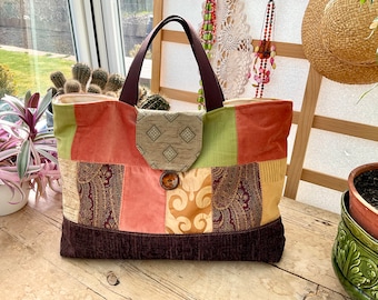 May include: A patchwork tote bag with a brown leather handle. The bag is made of different fabrics in shades of brown, green, pink, and beige. The bag has a large flap with a button closure.