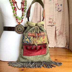 Könnte beinhalten: Eine grüne, braune und rote Patchwork-Kordeltasche mit einer bordeauxfarbenen Tasche und einem Fransenbesatz. Die Tasche hat verschiedene gestickte Details und Bommeln.