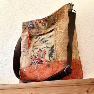 May include: A hobo-style shoulder bag featuring a patchwork design of floral tapestry fabric in shades of beige, red, and green, with a solid burnt orange textured base. It includes a dark brown adjustable strap, metal rivets, and a small orange tassel.