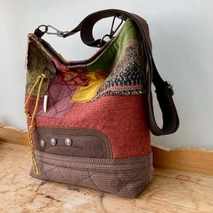 May include: A hobo-style shoulder bag featuring a patchwork design of various textured fabrics in shades of red, brown, green, and yellow. The bag includes a brown corduroy base, a dark brown leather-like accent panel with three metal studs, and a dangling white tusk-shaped charm on a silver chain.