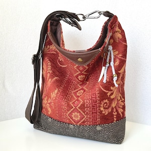 May include: A hobo bag featuring a red and orange textured fabric with a floral and geometric pattern. The base is dark grey with a snakeskin-like texture and metal studs. It includes a brown adjustable strap and a decorative white tassel charm.